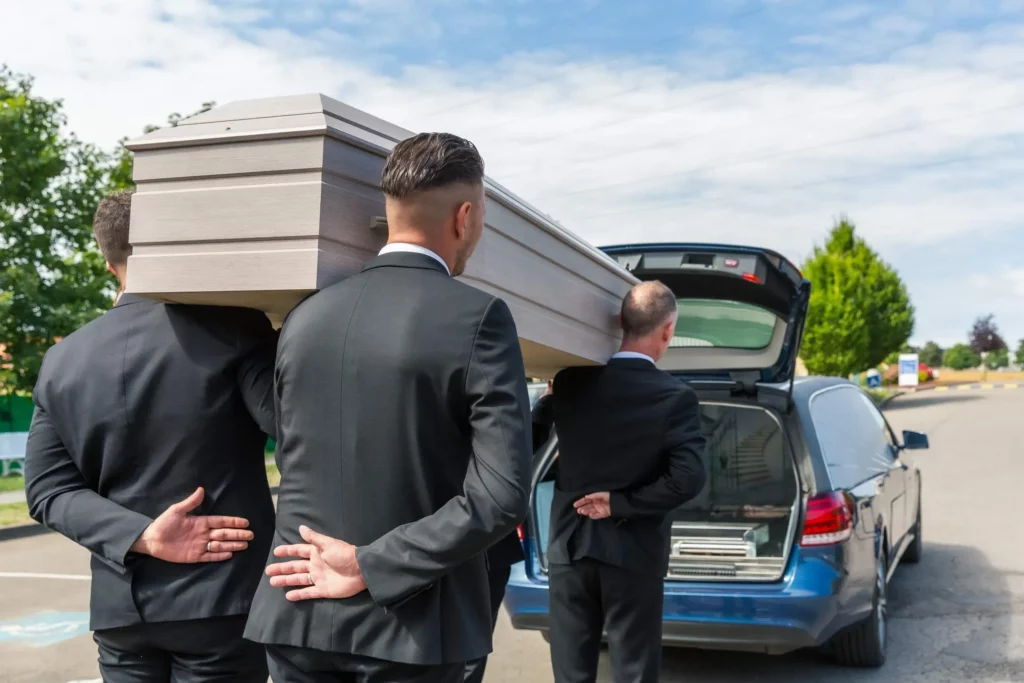 Porteurs funéraires en costume sombre portant un cercueil en bois clair sur leurs épaules.